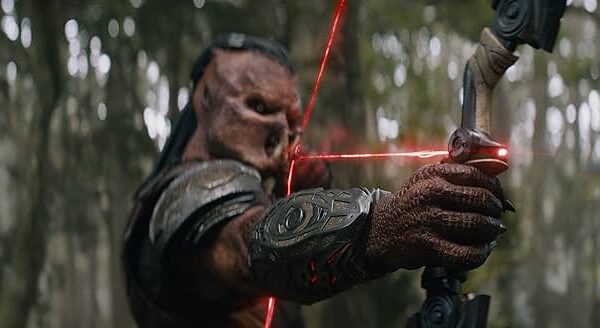 Depredador Tierras Salvajes — Estreno exclusivo en cines este noviembre Una criatura alienígena de Depredador Tierras Salvajes, vestida con armadura, apunta con un arco de alta tecnología con mira láser roja en un bosque. Su piel rugosa y texturizada y su intimidante rostro enmascarado lo convierten en un ser temible entre los árboles.